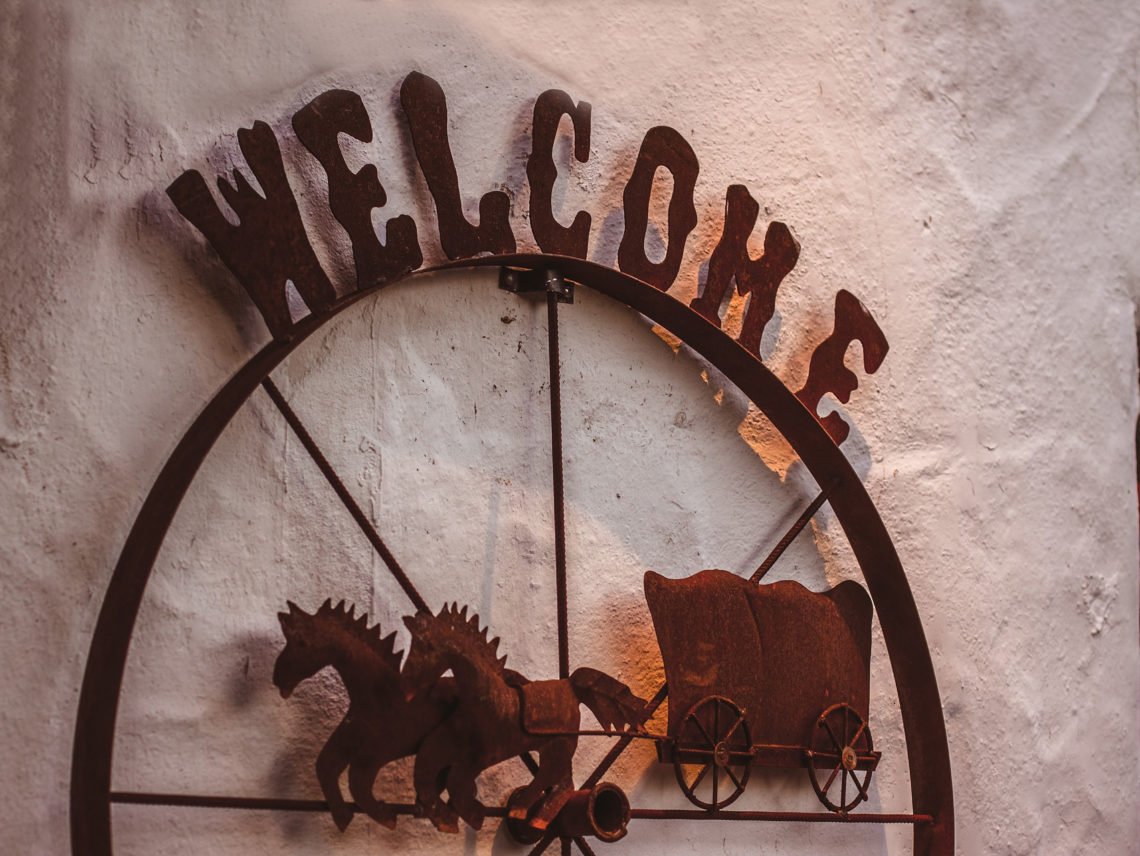 Rostig järnskylt på vitkalkad vägg med texten Welcome och en westernvagn dragen av två hästar