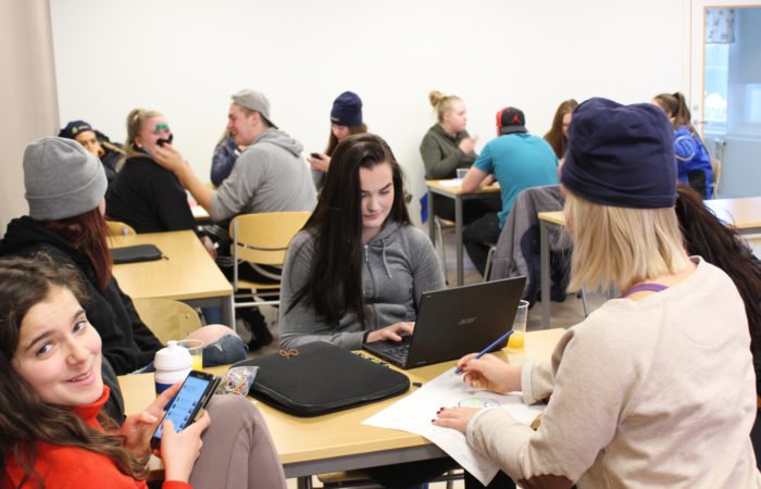 Elever i klassrum. En framför en dator, en upptagen med mobilen, en skriver på papper, medan två i bakgrunden leker med inspirationsmaterialet och skapar mustacher till varann.