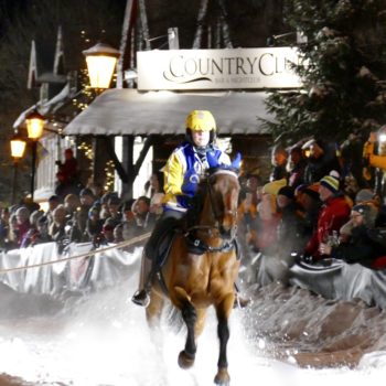 Varmblodshäst galopperar så snön ryker, ryttare bär Wångens gula och blå kläder. I bakgrunden syns publik till skijoring-tävling och skylt med texten Country Club.