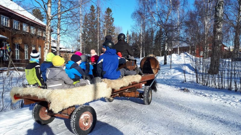 I en vagn täckt med fårskinn sitter en grupp barn samt kuskelev Anton. Två kuskar på kuskbocken. 