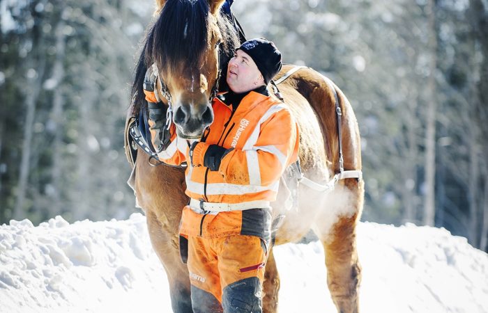 Man i varselkläder och mössa står bredvid och håller i sin häst i ett snöigt skogslandskap. Det är kallt, utandningsluften syns från hästen
