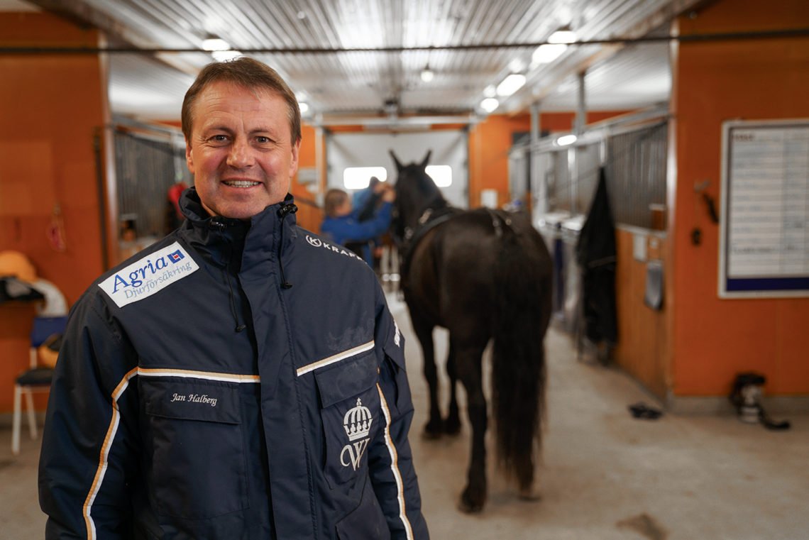 Jan Halberg tränare i stallet i mörkblått travställ med Wångens sigill på står i en stallgång under campkontroll. I bakgrunden skymtar en häst och en hästskötare.