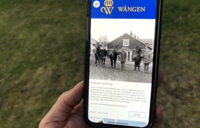 Guidad tur med vår app. Bilden föreställer en mobiltelefon och första sidan i appen med en svartvit bild där fyra män står med hästar framför ett av Wångens stall. Tidigt 1920-tal