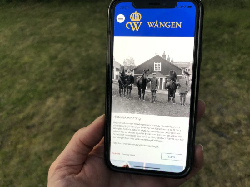 Guidad tur med vår app. Bilden föreställer en mobiltelefon och första sidan i appen med en svartvit bild där fyra män står med hästar framför ett av Wångens stall. Tidigt 1920-tal