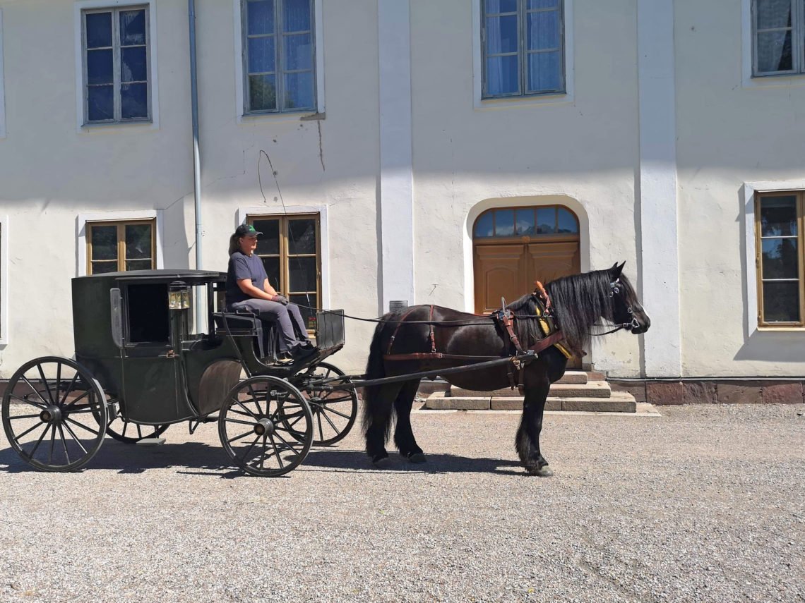 Svart häst spänd framför täckt vagn av klassisk modell står framför en herrgårdsliknande byggnad