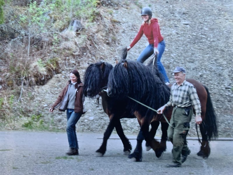 Kvinna och äldre man leder två nordsvenska brukshingstar med mycket man på en grusplan. På ryggen med ett ben på varje häst står en kvinna iförd jeans, röd tröja och ridhjälm. Med böjda knän håller hon balansen 