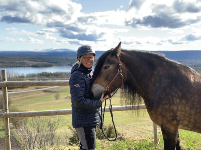 Kvinna iförd hjälm och jacka står och håller i en nordsvensk brukshäst. I bakgrunden syns en vidunderlig utsikt över sjö och fjäll. Det är vår.