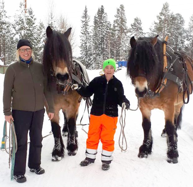 Två kvinnor bredvid varsin nordsvensk brukshäst i ett snöigt skogslandskap. 