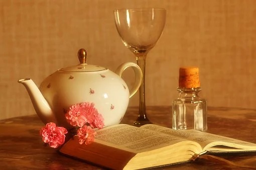 Fina loppis fynd på ett bord. En söt tekanna i porslin, ett glas på hög fot, en liten glasflaska med kork och en uppslagen bok.