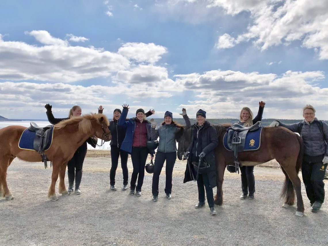Sex glada deltagare från hästskötarkurs islandshäst vinkar med händerna i luften. De står bredvid två sadlade islandshästar utomhus en solig vårdag