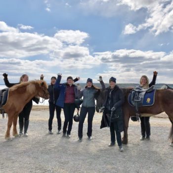 Sex glada deltagare från hästskötarkurs islandshäst vinkar med händerna i luften. De står bredvid två sadlade islandshästar utomhus en solig vårdag