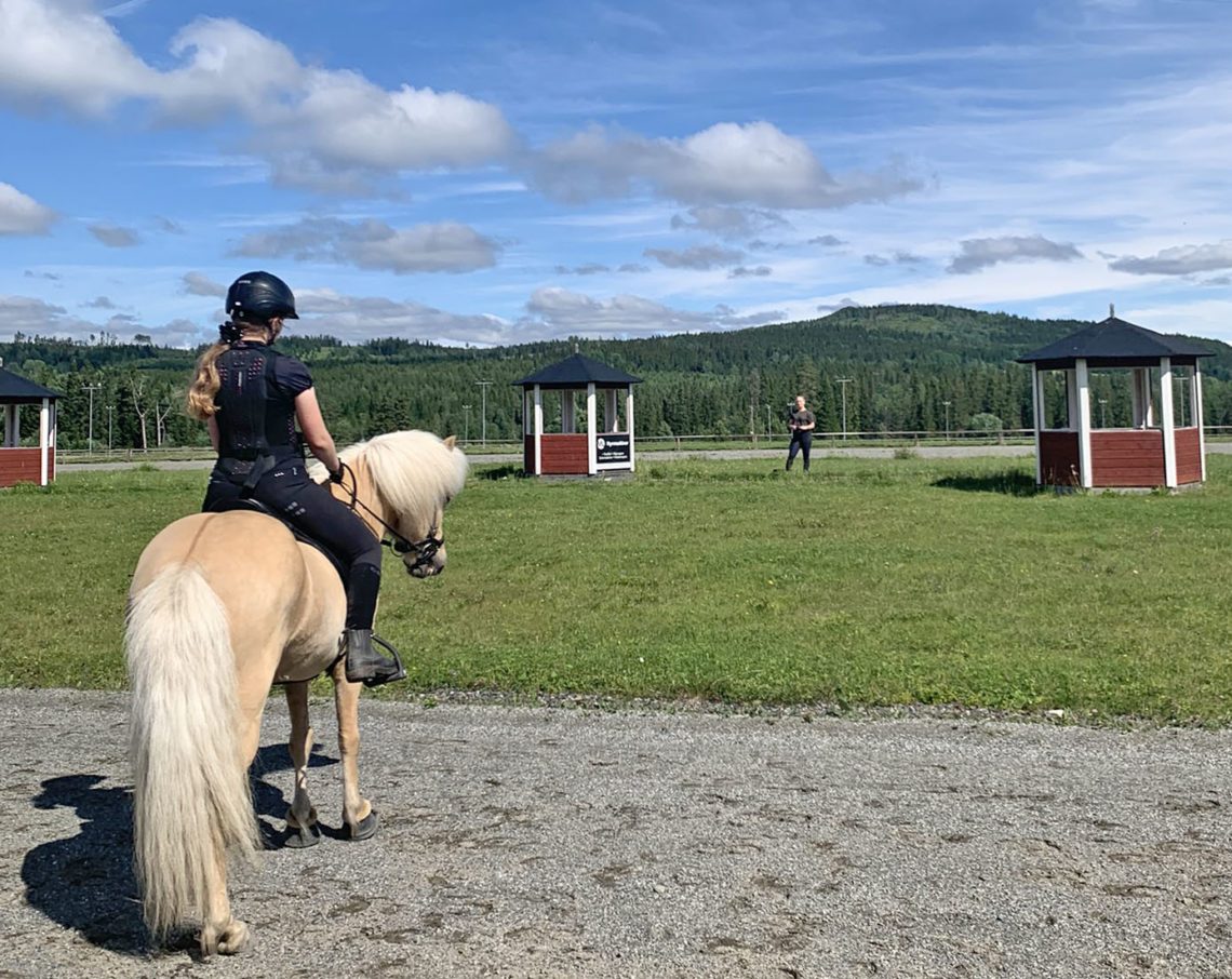 Flicka på ljusfuxfärgad islandshäst står vid ovalbana och tittar mot instruktören