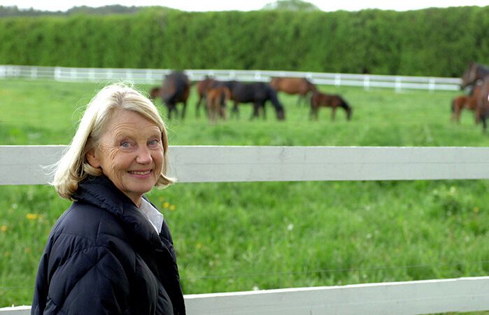 Margareta Wallenius-Kleberg står framför ett vitt staket och tittar in i kameran och ler. I bakgrunden syns en flock hästar på grönbete.