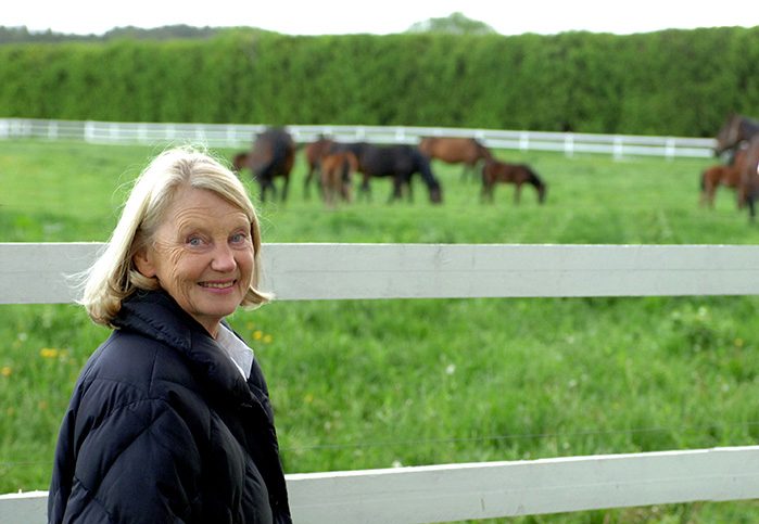 Margareta Wallenius-Kleberg står framför ett vitt staket och tittar in i kameran och ler. I bakgrunden syns en flock hästar på grönbete.