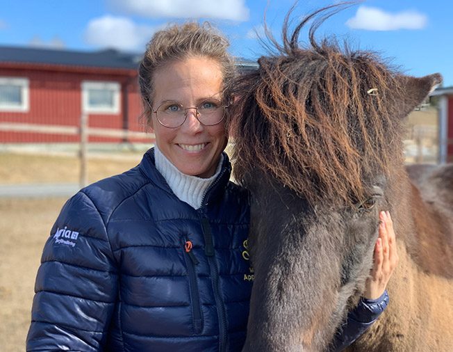 Malin Appelqvist står i en hage tillsammans med en islandshäst. Hon har en blå täckjacka med Wångens sigill på och ler in i kameran. Det är en soligt vårig dag.