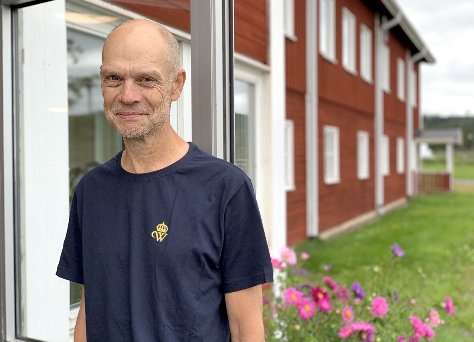 Man står och håller upp en dörr. Han har Wången-t-shirt på sig. Bakom syns en röd byggnad och gräsmatta och blommor