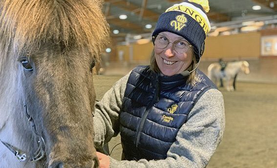 Camilla Hed står i ridhuset redo för preparandkurs! På bilden håller hon lektion för hippologstudenter i årskurs 3. På huvudet har hon en mössa med tofs med Wångens sigill. Hon har headset och ser glad ut. I bakgrunden syns en student rida en vit islandshäst.