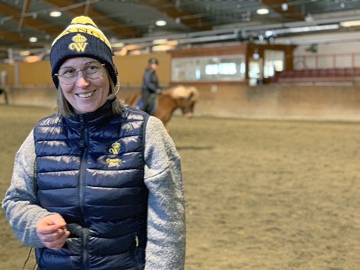 Camilla Hed står i ridhuset redo för preparandkurs! På bilden håller hon lektion för hippologstudenter i årskurs 3. På huvudet har hon en mössa med tofs med Wångens sigill. Hon har headset och ser glad ut. I bakgrunden syns en student rida en fuxfärgad islandshäst.