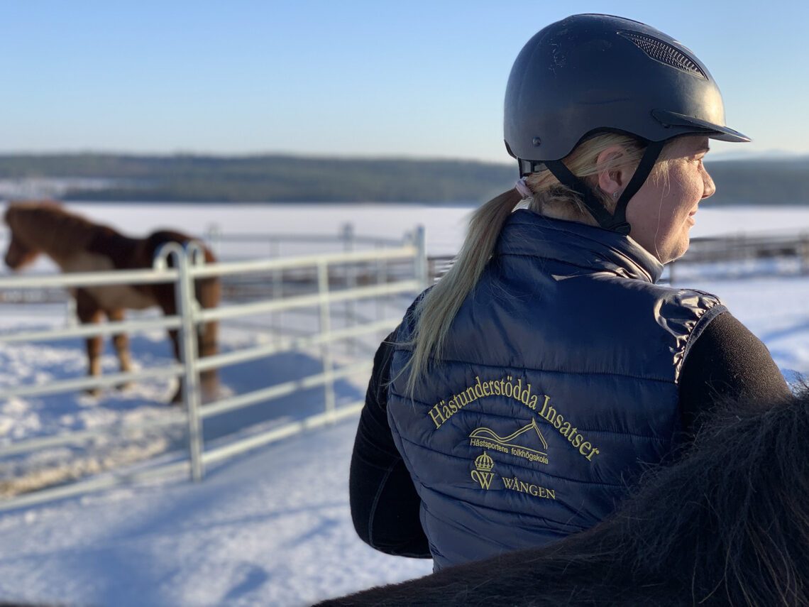 En solig vinterdag på Wången står en kvinna med hjälm och en väst med texten Hästunderstödda insatser broderat på ryggen. I bakgrunden syns en rundkorrall med en häst i.