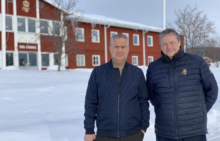 Ulf och Håkan står framför en stor röd byggnad med skylten Skol- och ridhus. Det är vinter och mycket snö på marken.