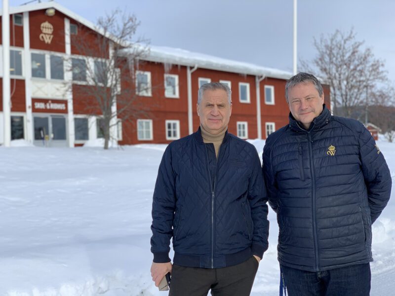 Ulf och Håkan står framför en stor röd byggnad med skylten Skol- och ridhus. Det är vinter och mycket snö på marken.