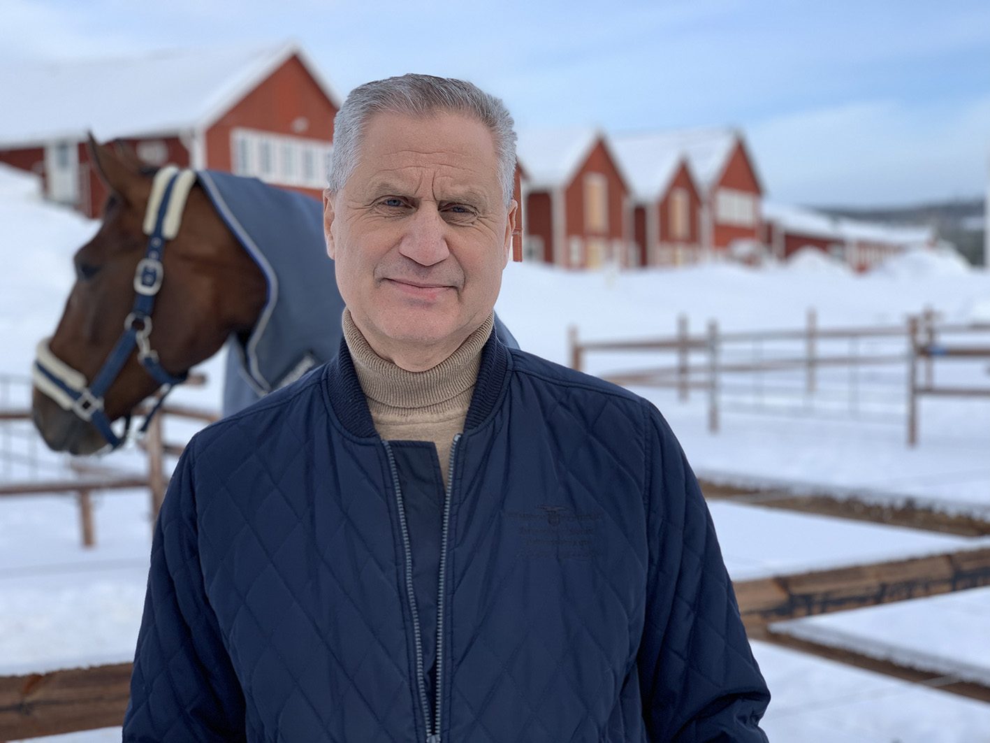 Ulf står framför en hage på Wången. Det är vinter och i bakgrunden syns huvudet på en varmblodstravare i täcke och grimma.