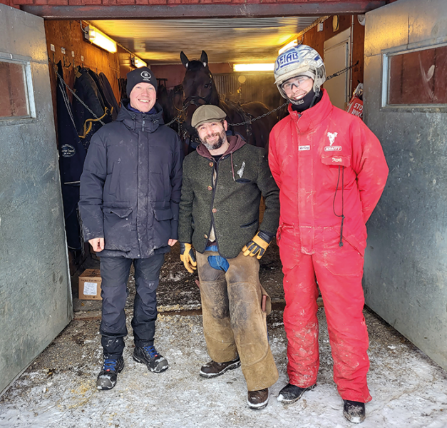 Tre män står i en stalldörr i vinterkläder. Inne i stallet syns en varmblodstravare uppställd på stallgången.