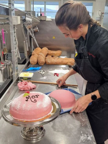 Kvinna står i ett industrikök och förbereder tårtor. De är rosa marsipantårtor med texten Mor på