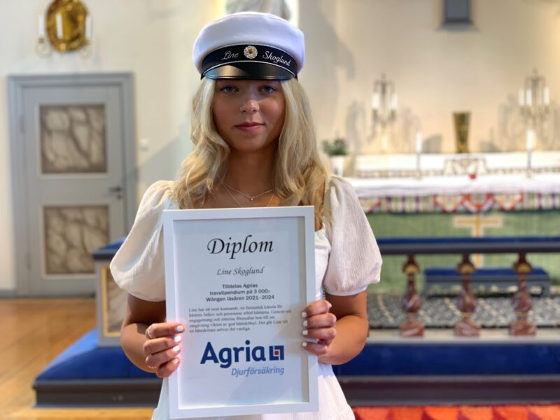 En ung kvinna i studentmössa håller upp ett stipendium med text Diplom och Agrias logoyp. Hon har vit klänning och står i kyrkan på avslutningsdagen. 