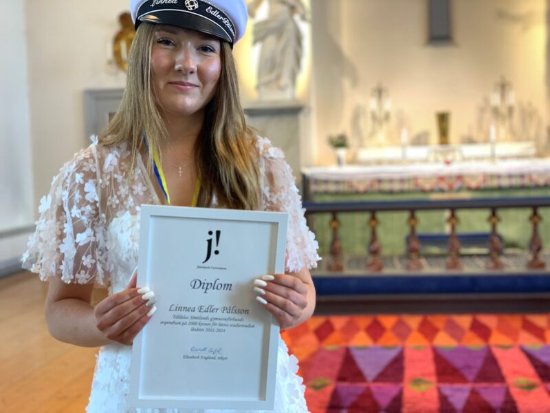 En ung kvinna i studentmössa håller upp ett stipendium. Hon står i kyrkan på avslutningsdagen.