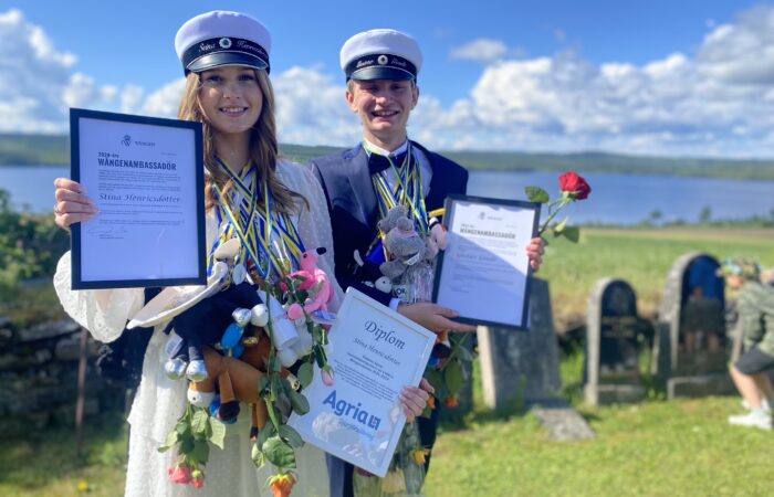 Stina och Gustav står finklädda med studentmössorna på runt halsen har de blågula band med blommor hängandes. I händerna håller de stipendierna. Det är en vacker sommardag. I bakgrunden syns Alsensjön.