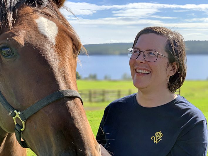 Kvinna i blå t-shirt med Wångens gula sigill ler och tittar på en varmblodstravare som hon leder i grimma. I bakgrunden syns hagar och en sjö.