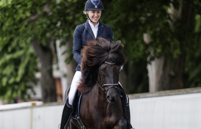 Madelen Johanson rider en svart islandshäst. Hon är tävlingsklädd i vita ridbyxor, stövlar och kavaj och ser glad ut. I bakgrunden syns en vit stenmur och höga björkar