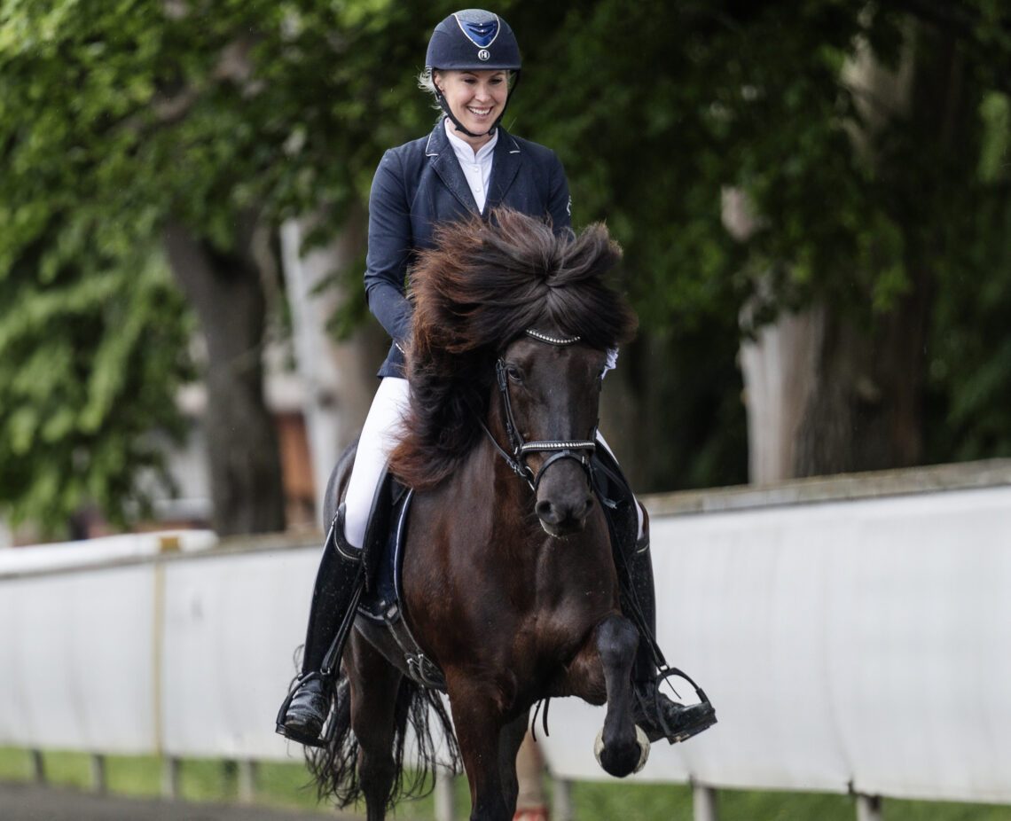 Madelen Johanson rider en svart islandshäst. Hon är tävlingsklädd i vita ridbyxor, stövlar och kavaj och ser glad ut. I bakgrunden syns en vit stenmur och höga björkar