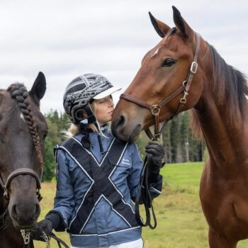 Flicka i kuskdress mellan två hästar blundar och ser ut att lukta på hästens mule