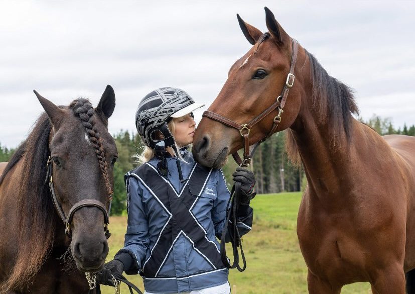 Flicka i kuskdress mellan två hästar blundar och ser ut att lukta på hästens mule